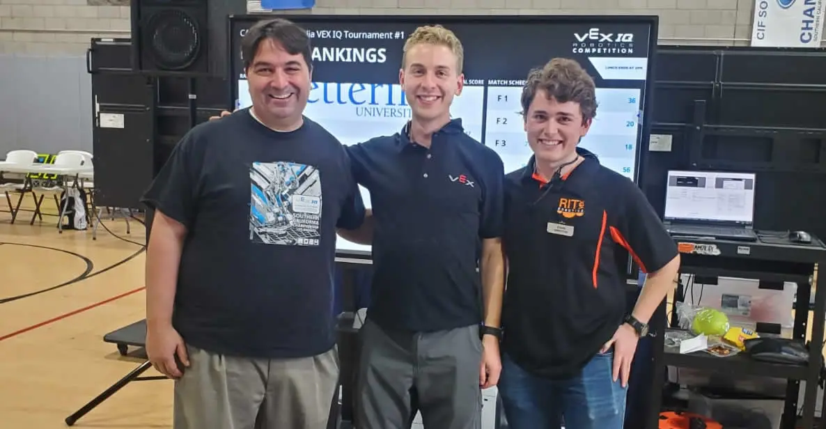 Three individuals, including Jeremy Sayers and Ben Lipper, posing at a VEX IQ robotics competition with rankings displayed in the background.