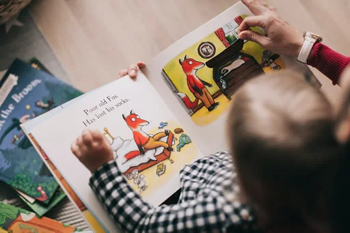 A child is about to start learning how to read. An adult reading a colorful children's book featuring a story about a fox. The book is open to a page with an illustration of a fox sitting on a bed and another fox near a fireplace. Other books are visible in the background.