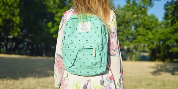 Back To School. A young girl wearing a floral dress and a light green backpack, standing outdoors with trees and a clear sky in the background.