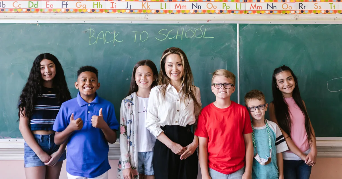 Teacher standing with six smiling students in front of a chalkboard that says 'Back to School.
