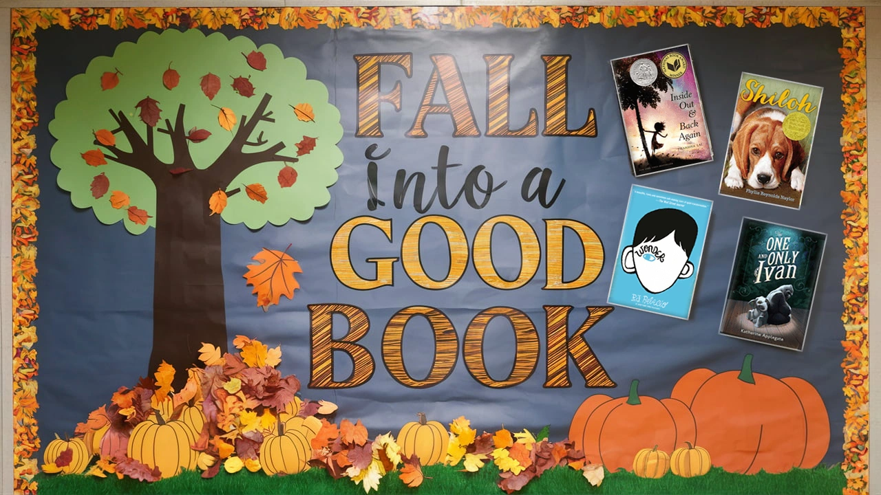 Bulletin board display with a fall theme titled 'Fall Into a Good Book,' featuring a large tree with autumn leaves, pumpkins, and images of book covers. The display is set against a blue background with a decorative fall-themed border.
