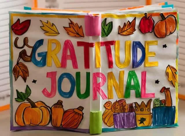 A colorful gratitude journal with the title "Gratitude Journal" written in large, bold letters across two pages. The cover is decorated with vibrant drawings of autumn leaves, pumpkins, and other seasonal elements. The journal pages are neatly held together with brightly colored tabs on the edges.