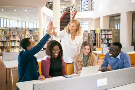 Parent Teacher Organization Success. A diverse group of five people sitting around a table in a library, collaborating on a project with a laptop. One person high-fives another while others smile and engage in the discussion. Bookshelves and large windows are visible in the background.